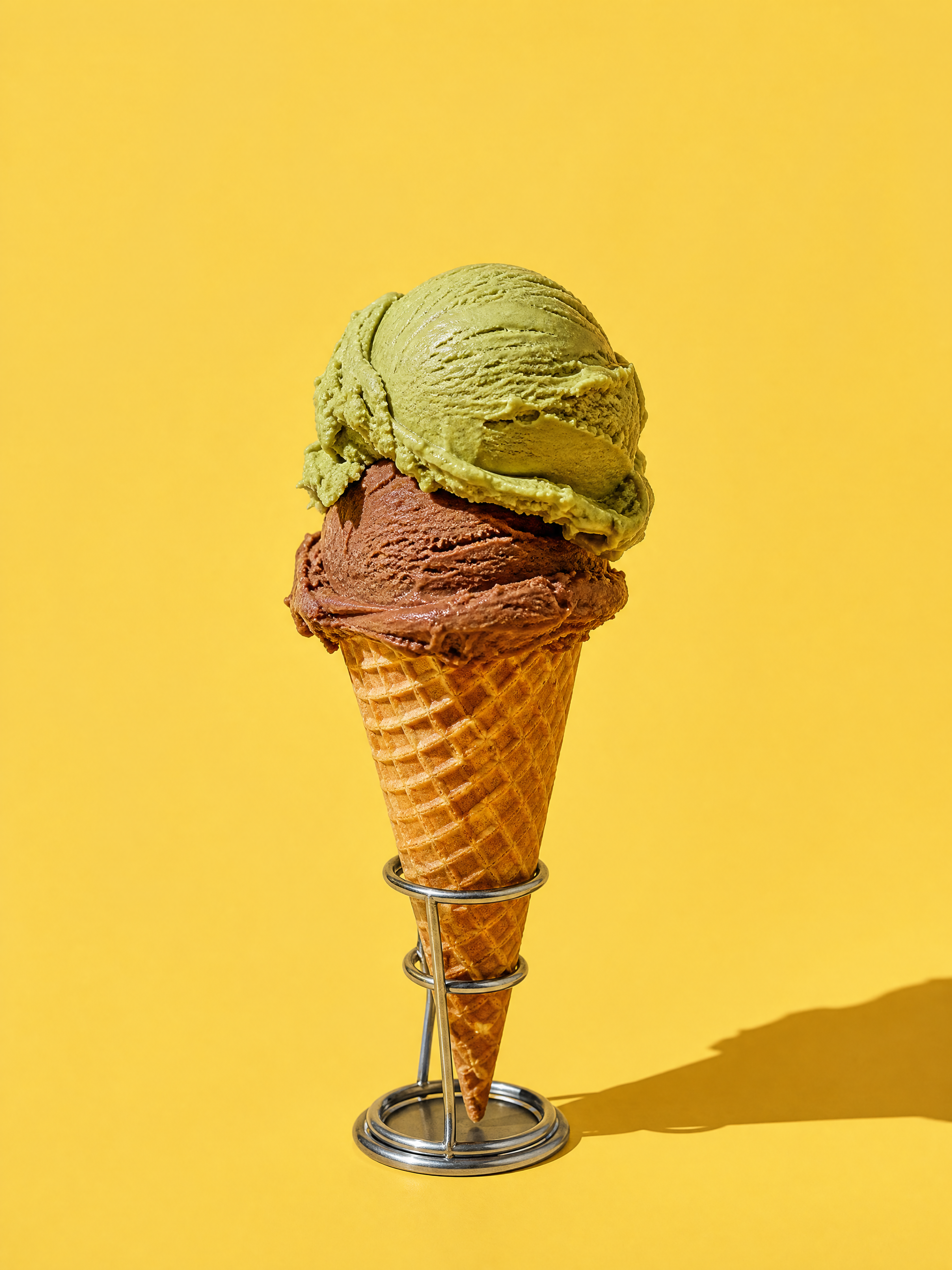 A waffle cone with a scoop of pistachio on a scoop of gianduia, standing in a metal holder against a vanilla-yellow wall